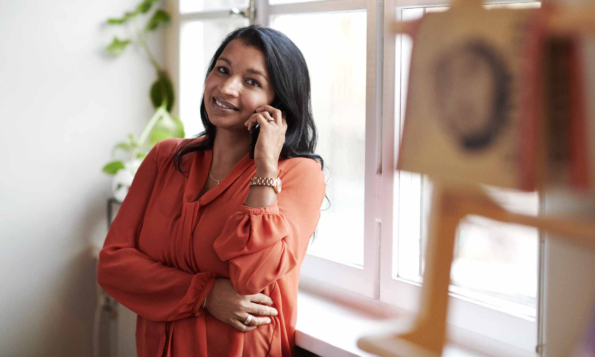 Leende affärskvinnan med orange blus talar i mobil vid ett fönster